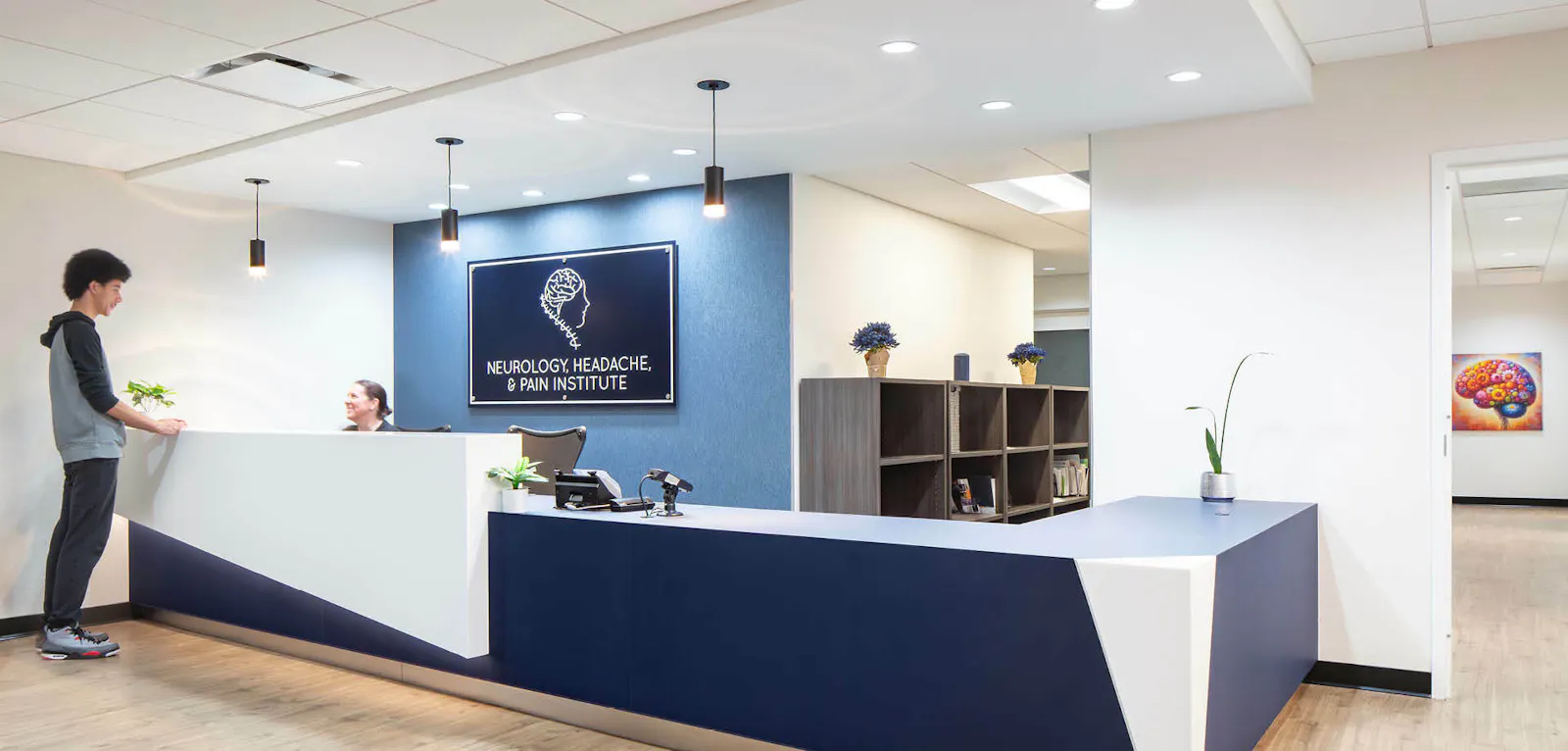 Two people speak at a sculptural blue and white reception desk for the Neurology, Headache & Pain Institute. 