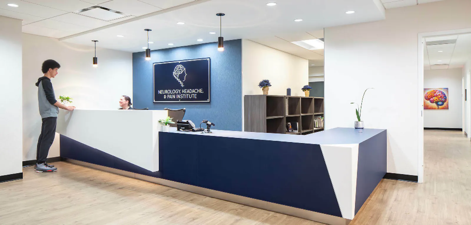 Two people speak at a sculptural blue and white reception desk for the Neurology, Headache & Pain Institute. 