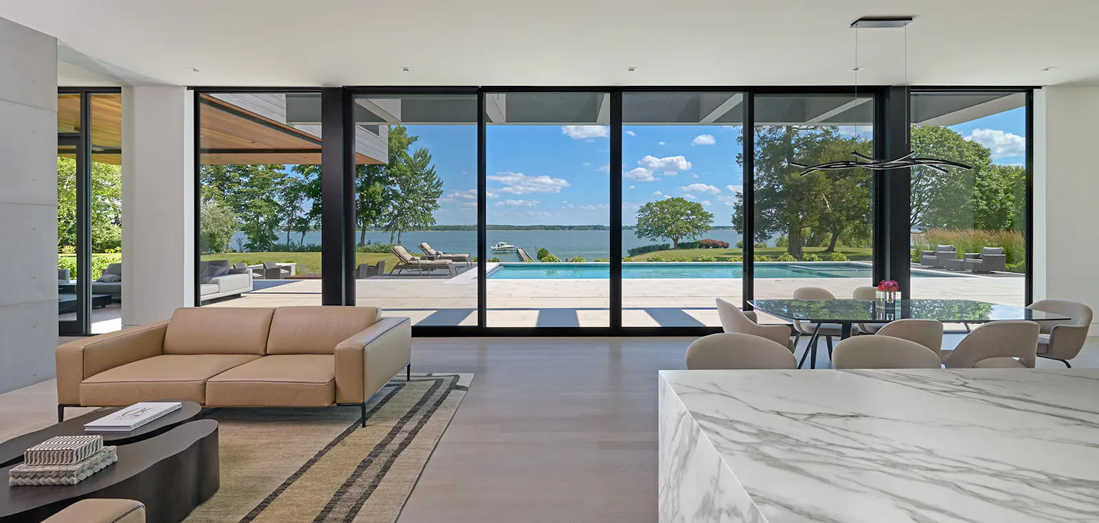 A minimal living room and dining room with neutral colored furniture is set against a wall of floor-to-ceiling windows overlooking a river. 