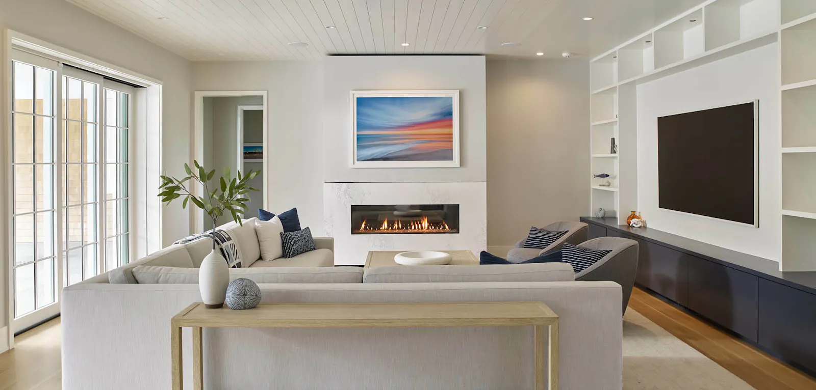 A calming living room includes a wall of built-in shelves and a statement fireplace, surrounded by luxurious white furniture. 