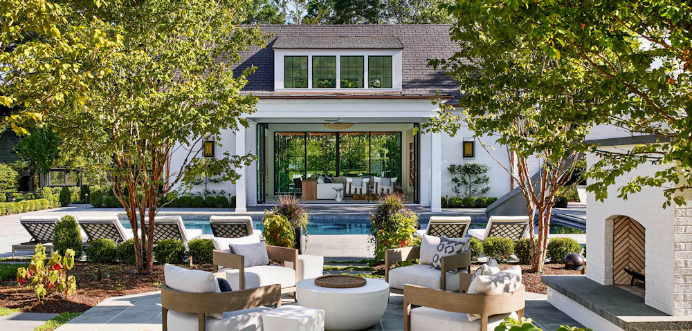 A luxurious double-sided poolhouse is seen behind a pool and outdoor fireplace surrounded by four chairs.